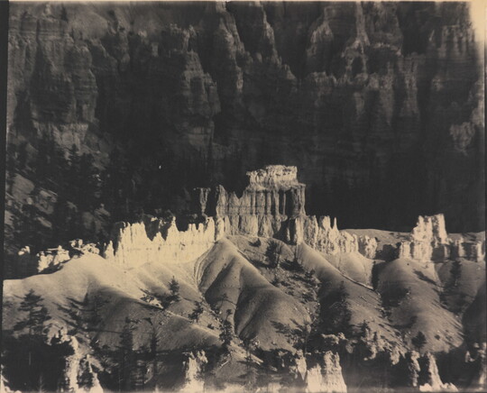 A black-and-white photograph of a landscape with hoodoos, or vertical spires of rock, on top of sand dune shapes with cliffs in the background.