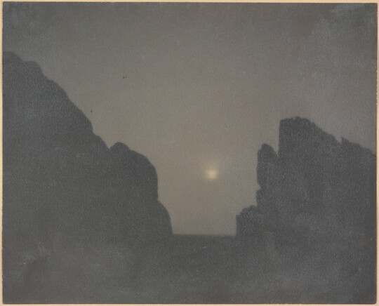 A black-and-white photograph of two rock formations in silhouette with a hazy moon in between them.