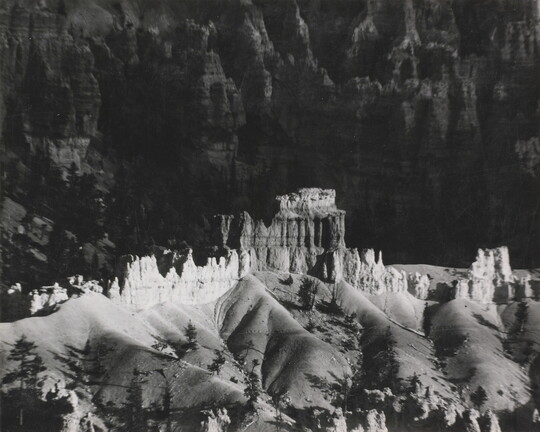 A black-and-white photograph of a landscape with hoodoos, or vertical spires of rock, on top of sand dune shapes with cliffs in the background.
