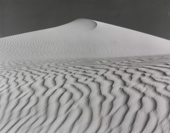 A sepia-toned photograph of a deeply-rippled sand dune rising to a point into the sky.