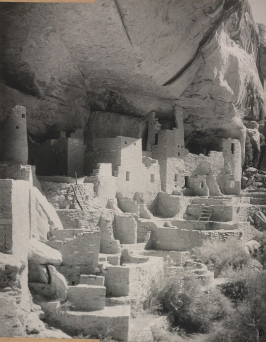 A black-and-white photograph of an abandoned, crumbling rock city built into the side of a desert cliff.