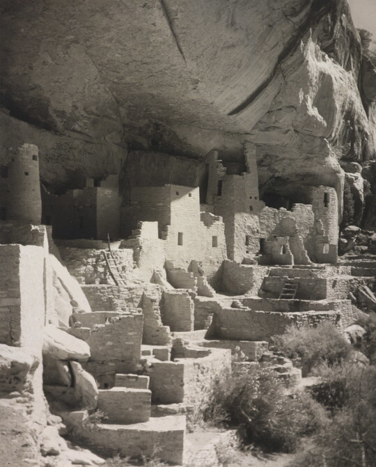 A black-and-white photograph of an abandoned, crumbling rock city built into the side of a desert cliff.