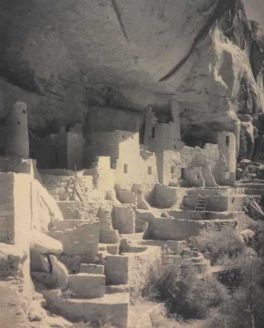 A black-and-white photograph of an abandoned, crumbling rock city built into the side of a desert cliff.