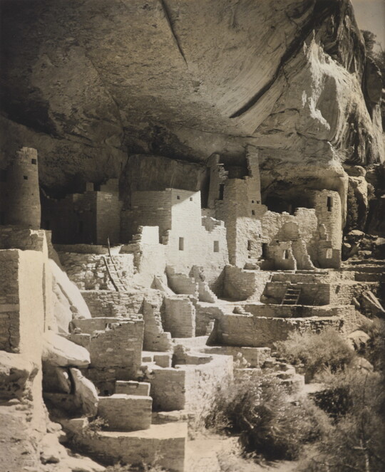 A black-and-white photograph of an abandoned, crumbling rock city built into the side of a desert cliff.