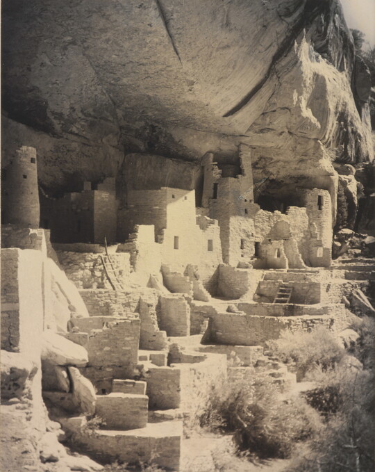 A black-and-white photograph of an abandoned, crumbling rock city built into the side of a desert cliff.