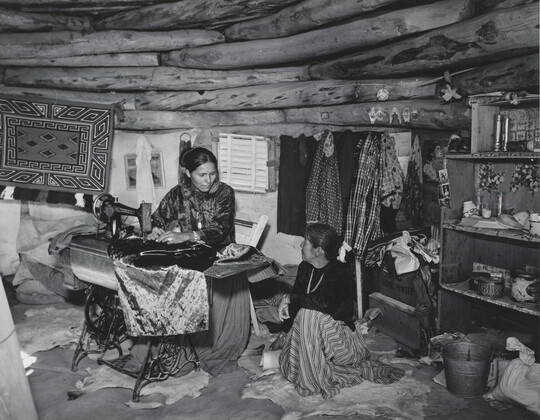 A black-and-white photograph of two Indigenous women seated, talking, and sewing inside of a low-ceilinged, wood structure.