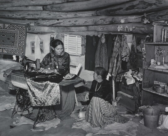 A black-and-white photograph of two Indigenous women seated, talking, and sewing inside of a low-ceilinged, wood structure.