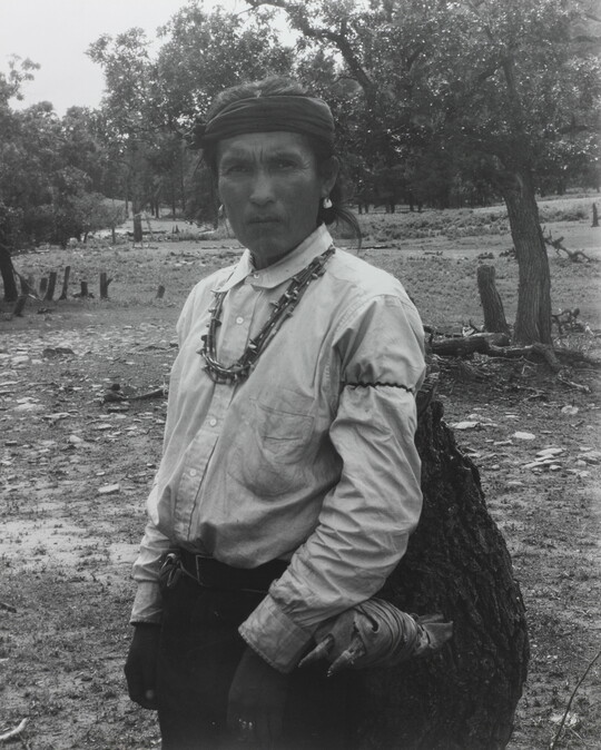 A black-and-white photograph of an Indigenous man carrying a large bag.