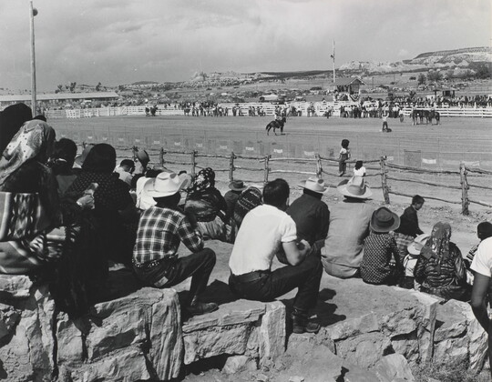 A black-and-white photograph of a group of spectators watching cowboys and horses in an outdoor arena.