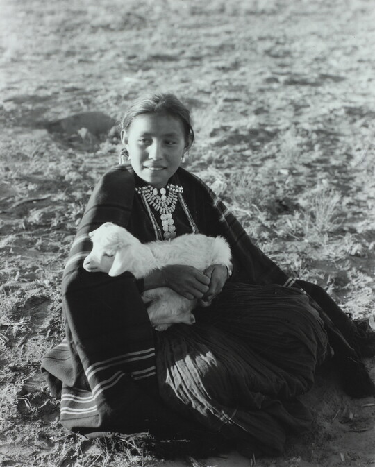A black-and-white photograph of an Indigenous girl sitting on the ground while holding a lamb.