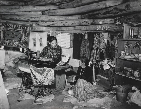 A black-and-white photograph of two Indigenous women seated, talking, and sewing inside of a low-ceilinged, wood structure.