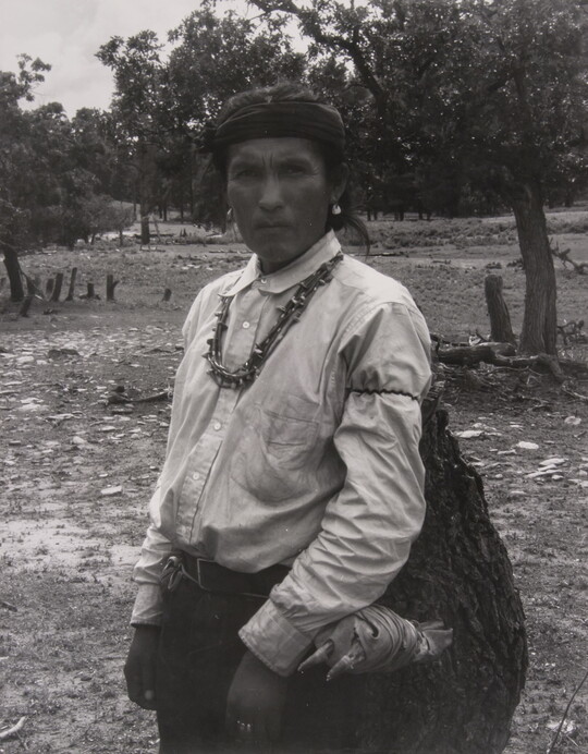 A black-and-white photograph of an Indigenous man carrying a large bag.