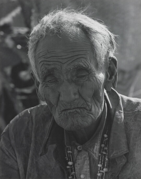 A black-and-white portrait photograph of an elderly Indigenous man with deep wrinkles and sunken cheeks.