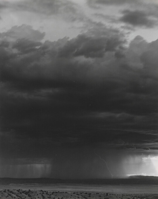 A black-and-white photograph of a dark storm cloud, heavy rain, and lightening over a desert.