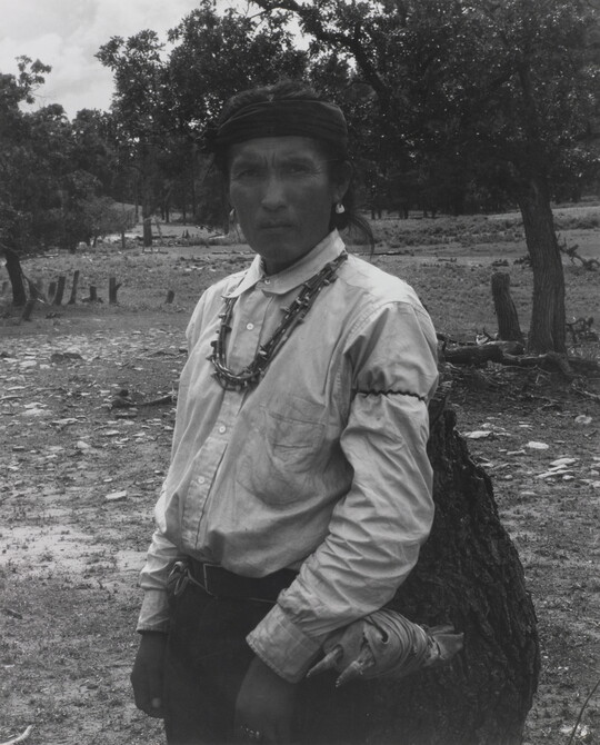 A black-and-white photograph of an Indigenous man carrying a large bag.