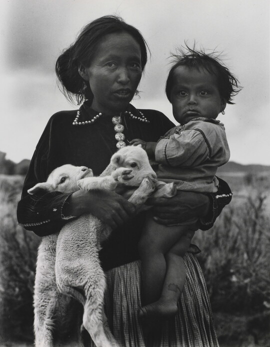 A black-and-white portrait photograph of a Native American woman holding a toddler on one hip and two lambs in her other arm.