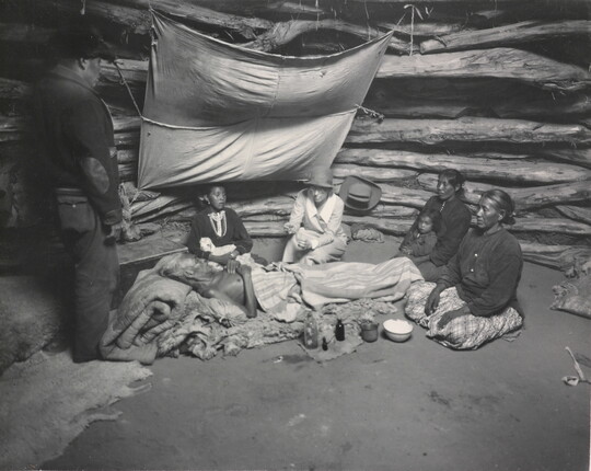 A black-and-white photograph of an Indigenous man laying on a pallet surrounded by other Indigenous people as a White woman holds a glass jar out to him.