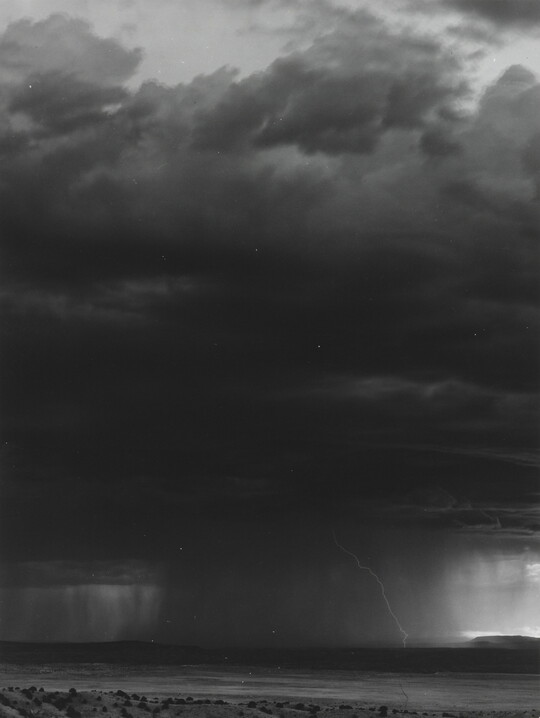 A black-and-white photograph of a dark storm cloud, heavy rain, and lightening over a desert.