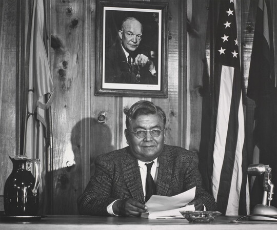 A black-and-white photograph of an Indigenous man dressed in a suit, seated at a desk between two flags and under a portrait of President Eisenhower.