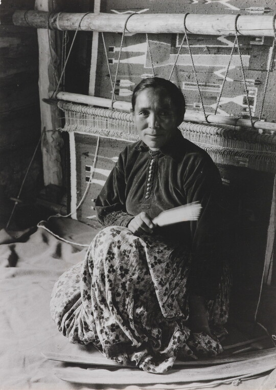 A black-and-white photographic portrait of a Native American woman sitting on the floor in front of a large loom.