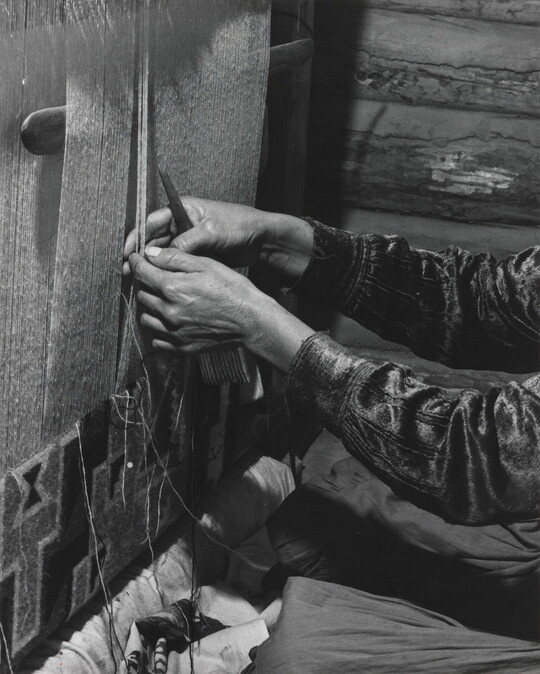 A black-and-white photograph of a person's hands weaving a design into a tapestry on a loom.