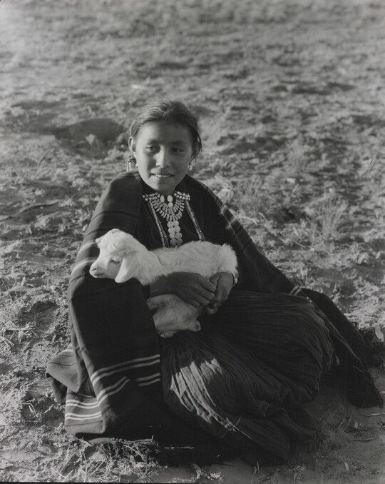 A black-and-white photograph of an Indigenous girl sitting on the ground while holding a lamb.