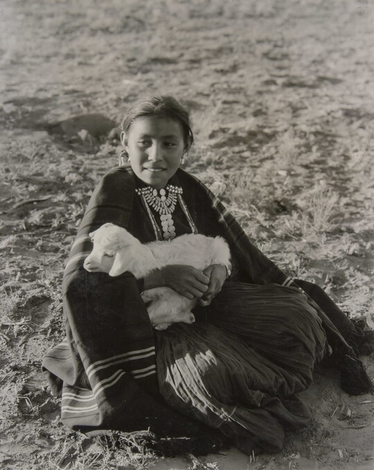 A black-and-white photograph of an Indigenous girl sitting on the ground while holding a lamb.
