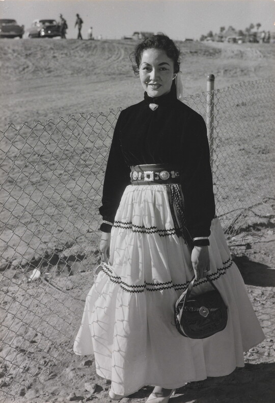 A black-and-white photograph of a young Indigenous woman wearing a long white skirt and dark blouse and standing in front of a chain link fence.