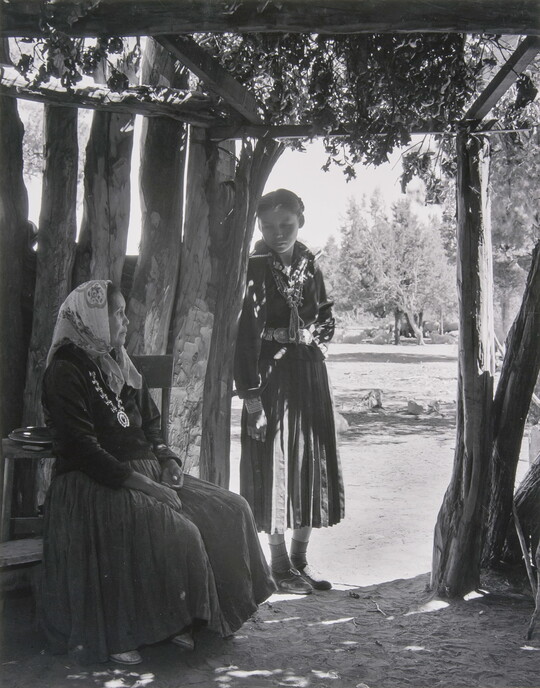 A black-and-white photograph of two Indigenous women, one seated and one standing, in a wooden structure open to outside.