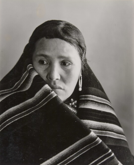 A black-and-white portrait photograph of an Indigenous woman with a blanket wrapped around her neck and shoulders.
