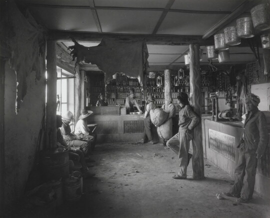 A black-and-white photograph of Indigenous people in a shop with a variety of products such as animal hides, tin pails, and lanterns hanging from the ceiling.