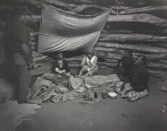 A black-and-white photograph of an Indigenous man laying on a pallet surrounded by other Indigenous people as a White woman holds a glass jar out to him.