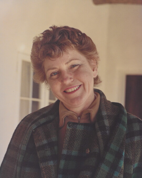 A color portrait photograph of a smiling, fair-skinned woman with red hair, wearing a plaid jacket, and standing on a porch.