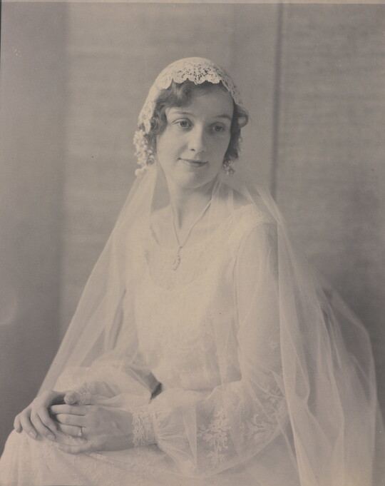 A black-and-white portrait photograph of a seated young White woman, hands folded on her lap, wearing a wedding dress with a long veil.