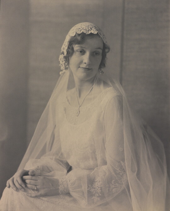 A black-and-white portrait photograph of a seated young White woman, hands folded on her lap, wearing a wedding dress with a long veil.