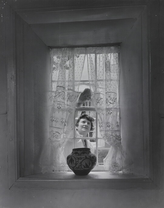A black-and-white photograph of a sheer curtained window with a Native American-style pot on the sill as a White woman cleans the window from outside.