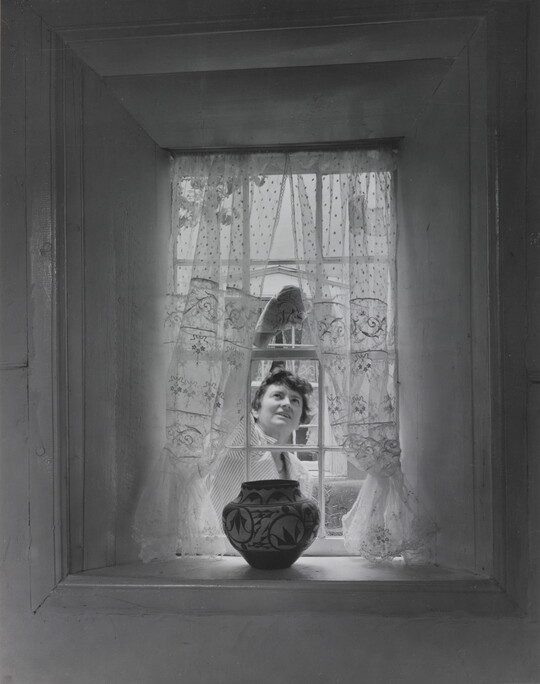 A black-and-white photograph of a sheer curtained window with a Native American-style pot on the sill as a White woman cleans the window from outside.