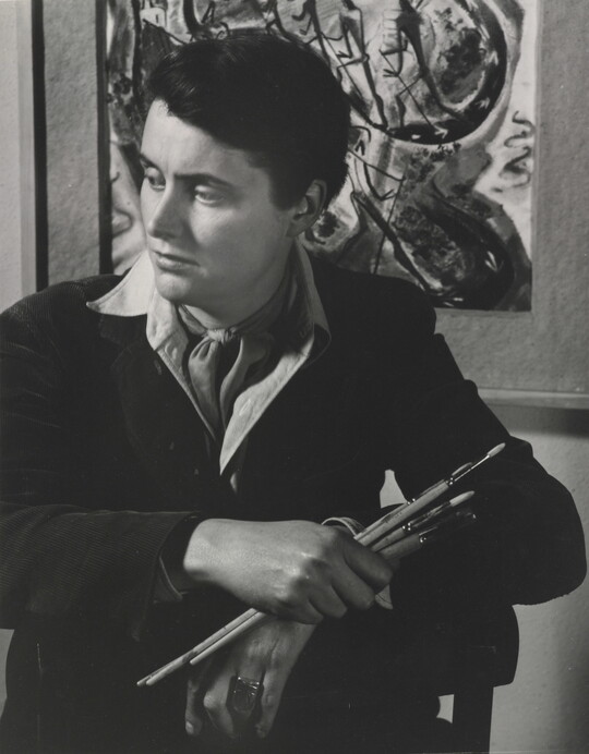 A black-and-white portrait photograph of a seated White woman holding paintbrushes in front of an abstract painting.