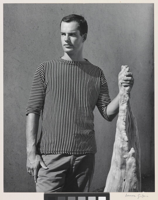 A black-and-white photograph of a White man with wearing a shirt with vertical stripes and holding a wooden object.