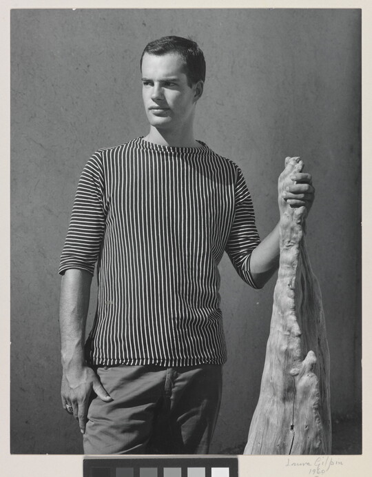 A black-and-white photograph of a White man with wearing a shirt with vertical stripes and holding a wooden object.