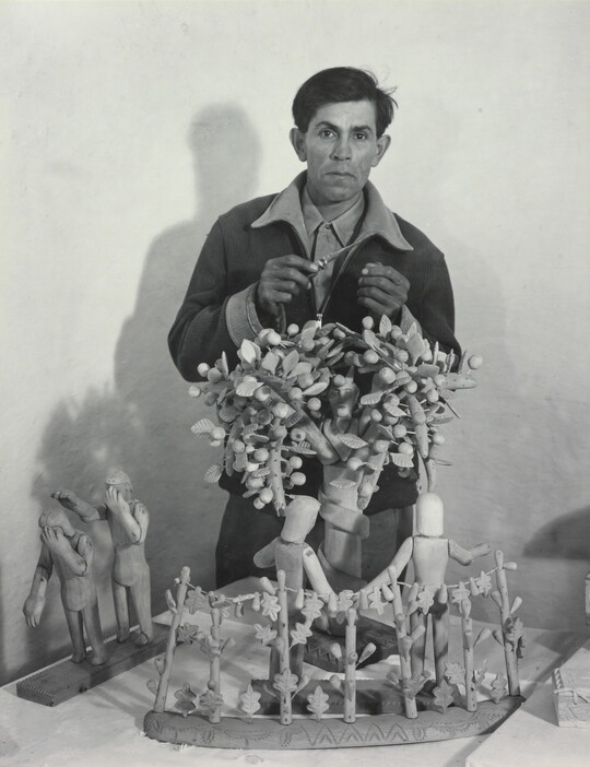 A black-and-white photograph of a medium-skinned man standing behind a wood sculpture of trees and human figures.
