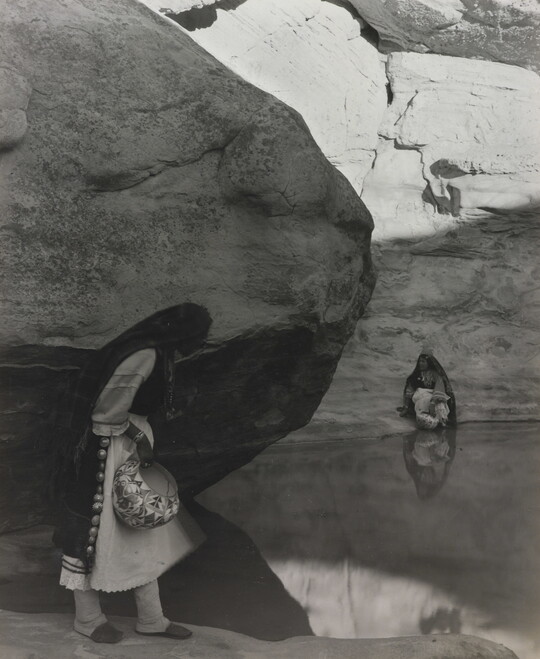 A black-and-white photograph of a woman with a long headscarf standing at the edge of a water hole surrounded by boulders and rock walls.