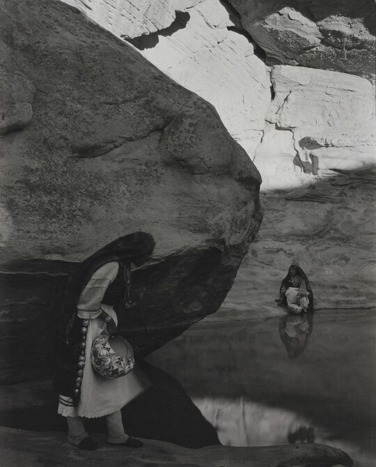 A black-and-white photograph of a woman with a long headscarf standing at the edge of a water hole surrounded by boulders and rock walls.
