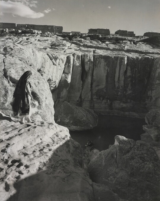 A black-and-white photograph of a woman with a long headscarf on a cliff, overlooking a water hole at the bottom.