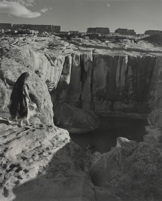 A black-and-white photograph of a woman with a long headscarf on a cliff, overlooking a water hole at the bottom.