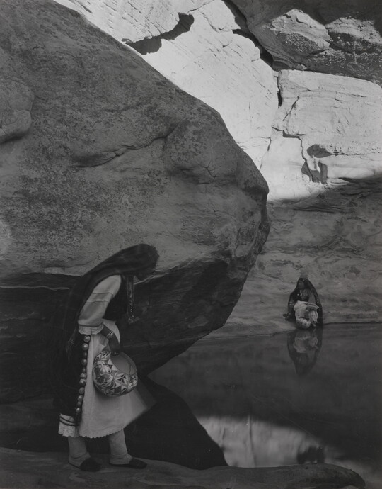 A black-and-white photograph of a woman with a long headscarf standing at the edge of a water hole surrounded by boulders and rock walls.