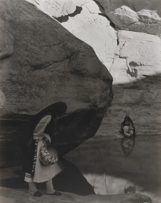 A black-and-white photograph of a woman with a long headscarf standing at the edge of a water hole surrounded by boulders and rock walls.