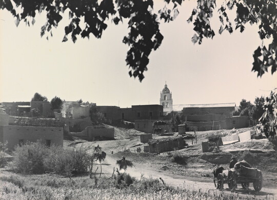 A black-and-white photograph of a town with adobe buildings and wagons and horses traveling on a dusty road.