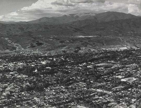 A black-and-white aerial photograph of a town in a mountain valley beneath puffy white clouds.
