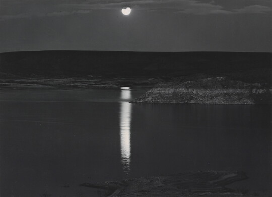 A black-and-white photograph of a night sky and full moon reflected across calm water.